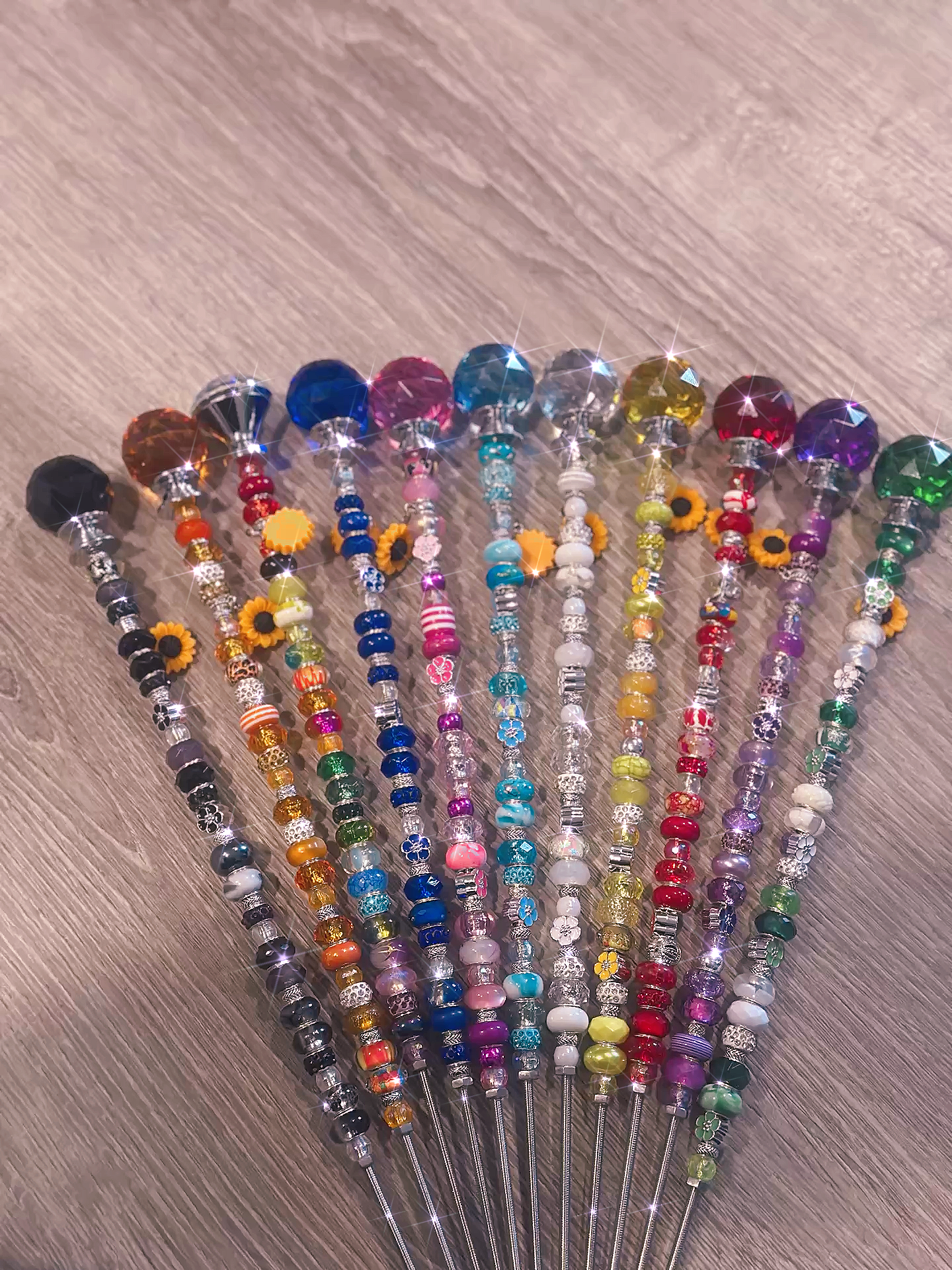 Collection of vibrant Garden Fairy Wands lined up on a wooden surface. Each handcrafted wand features colorful beads and charm details, ideal as a whimsical gift.