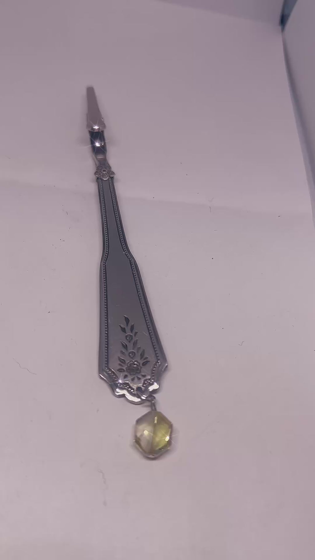 Handmade silver bracelet with a textured interlocking design and a dangling charm of green and transparent beads, displayed on a dark wood surface with warm ambient lighting.