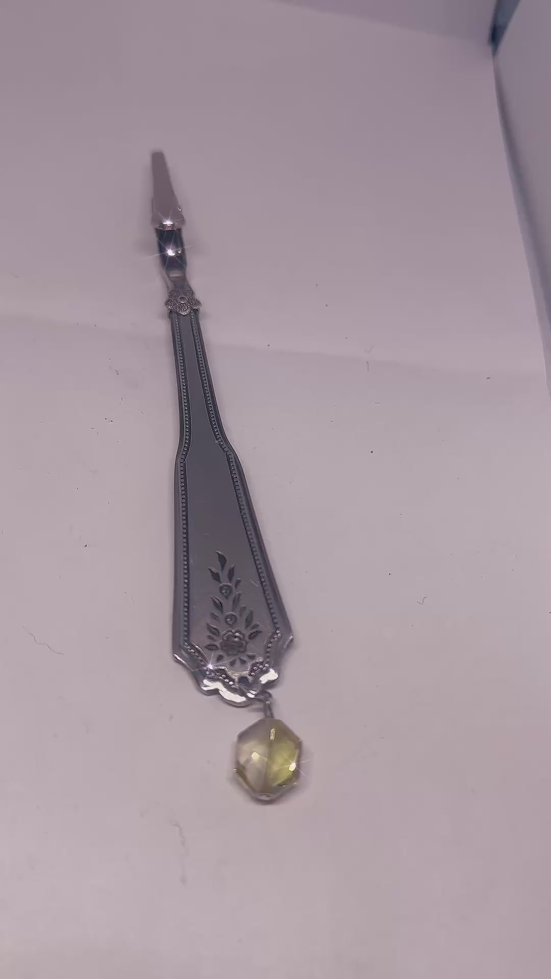 Handmade silver bracelet with a textured interlocking design and a dangling charm of green and transparent beads, displayed on a dark wood surface with warm ambient lighting.